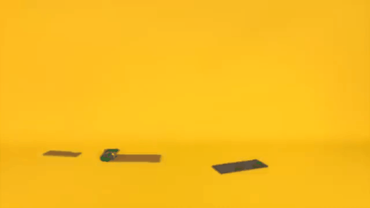 A yellow background with three objects on the surface: a small black object, a green and black stapler, and two black rectangular pieces.