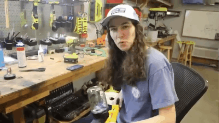 Emily The Engineer, with long brown hair wearing a gray baseball cap and gray t-shirt sitting at a cluttered workbench in a workshop, surrounded by tools and materials.