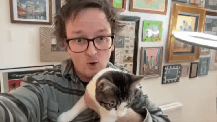 Half-Asleep Chris, taking a selfie while holding a cat in his arms in a room decorated with various framed pictures.