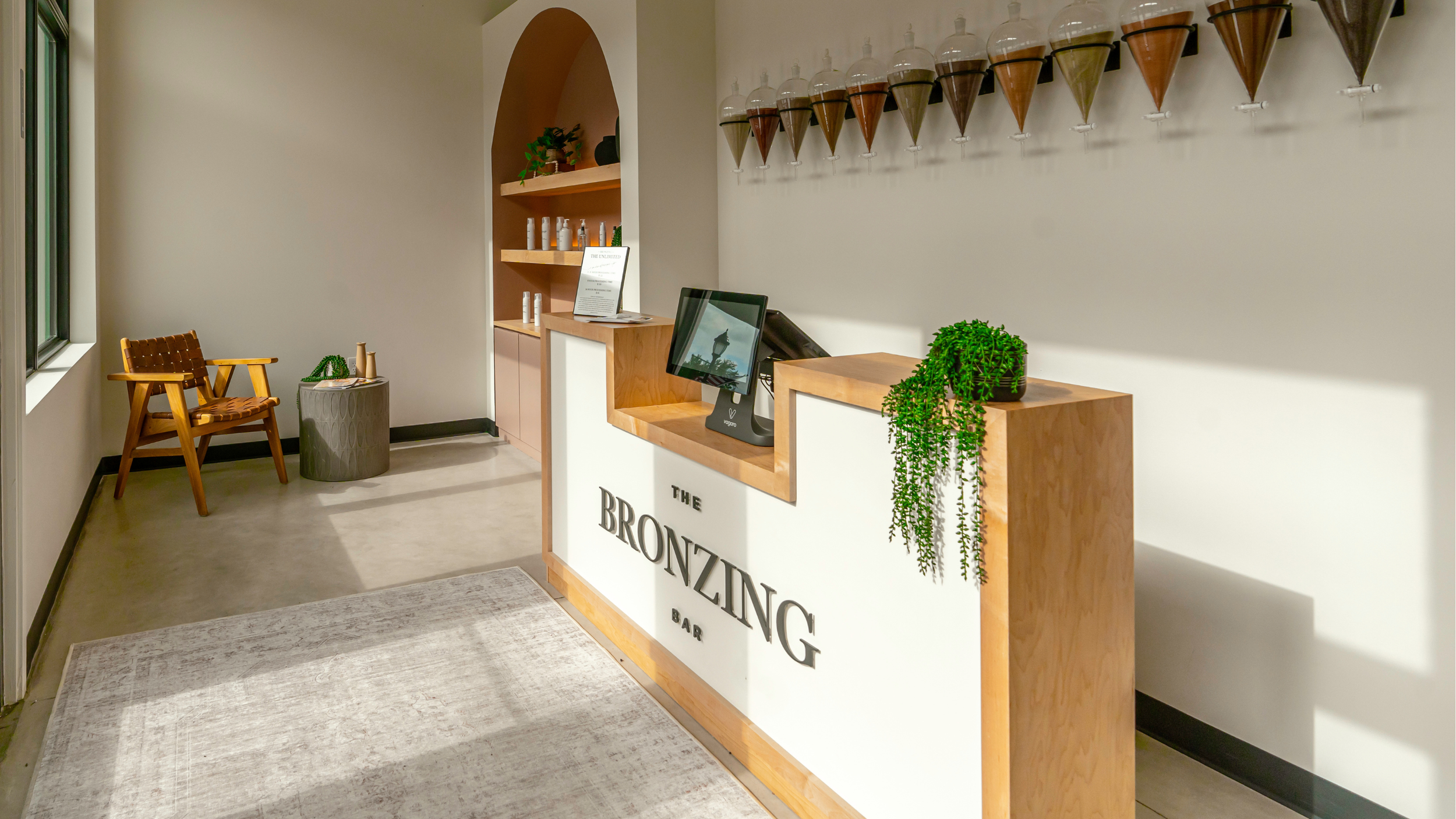 Reception area of a bar named The Bronzing Bar with a wooden counter, a computer, and green plants, a wall-mounted display of cone-shaped spice containers, wooden chairs, and shelves with bottles and decor.