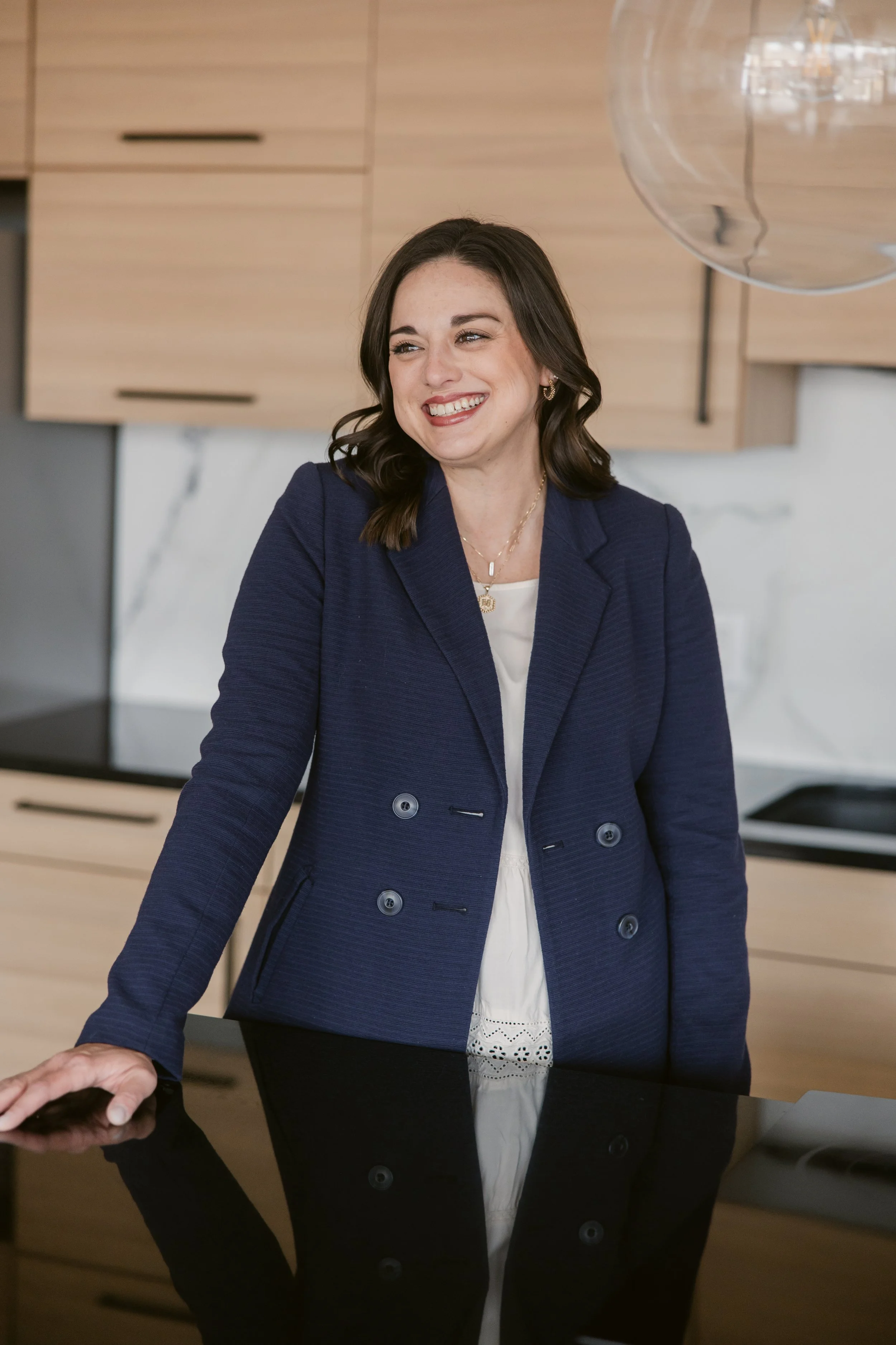 A woman with dark brown hair smiling and wearing a navy blue blazer, standing in a modern kitchen.