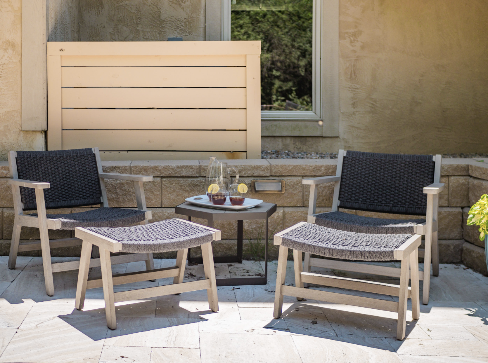 Two patio chairs with matching ottomans and a small table holding a pitcher and two glasses on a stone patio.