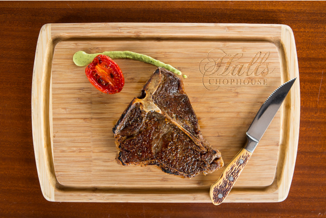 T-bone steak, roasted tomato, and a streak of herb sauce on a branded wooden serving board with a steak knife.
