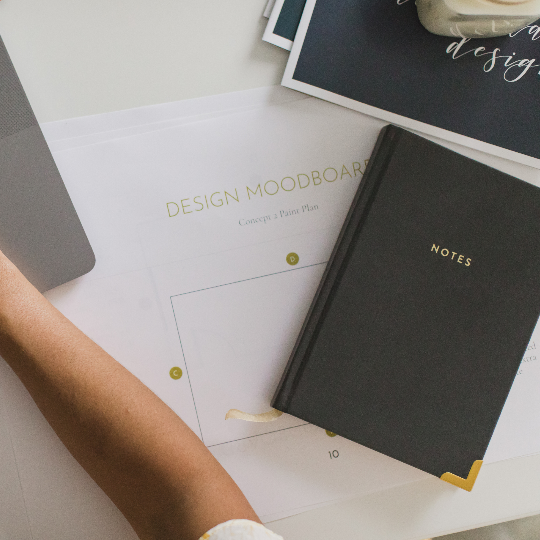 Top view of various design notebooks and mood boards on a white surface, with a person's arm and hand visible at the bottom.