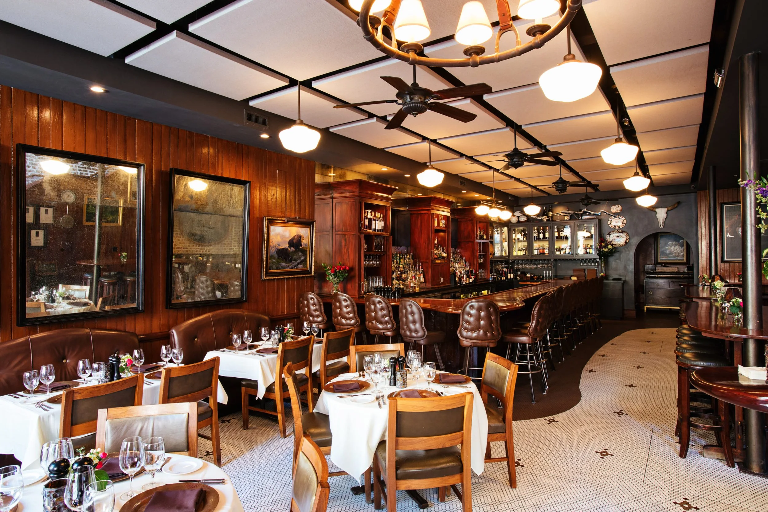 A restaurant dining room with white-clothed tables, leather booths, and a wooden bar with stools.