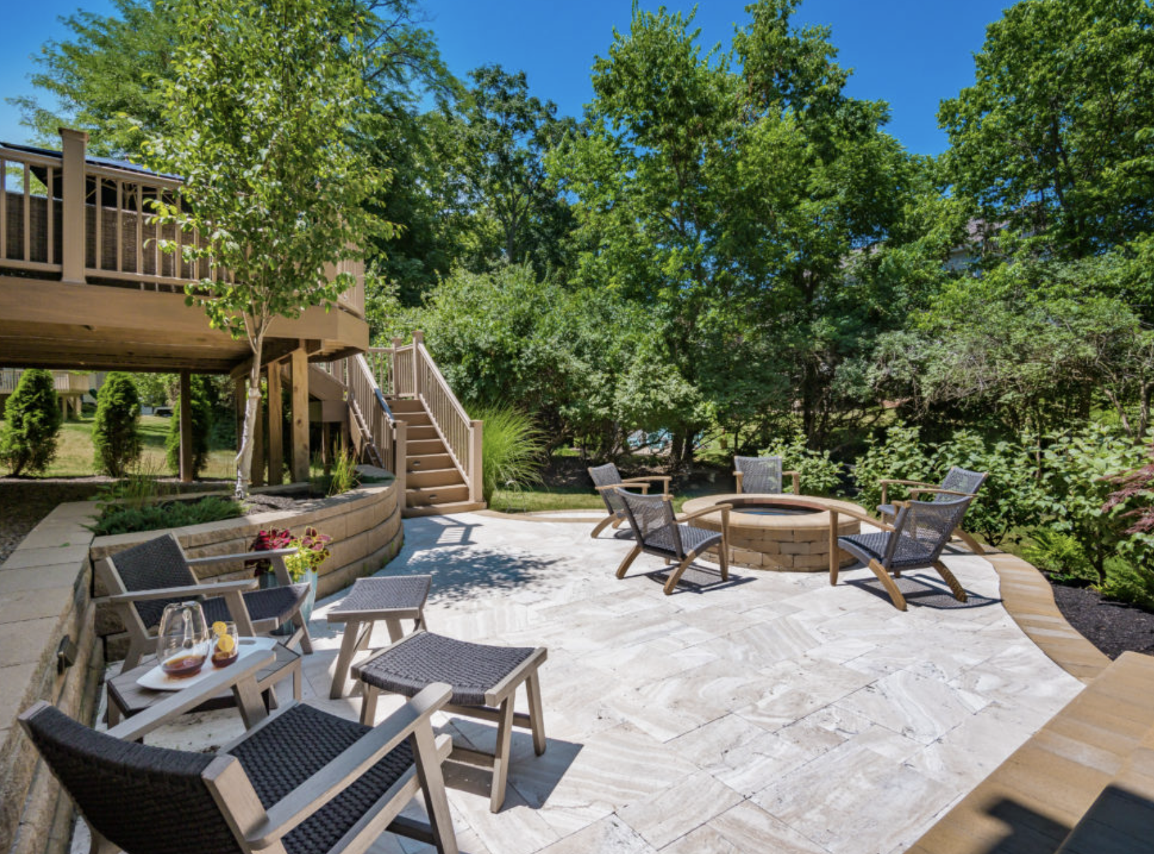 Backyard patio with stone flooring, a circular fire pit, outdoor chairs, and stairs leading to an elevated deck.