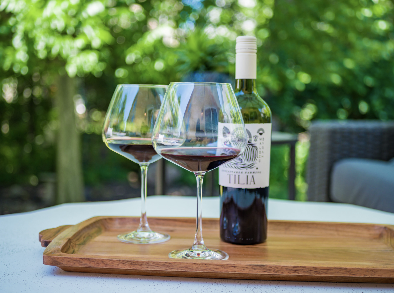 A bottle of Tilia red wine and two glasses of wine on a wooden tray outdoors.