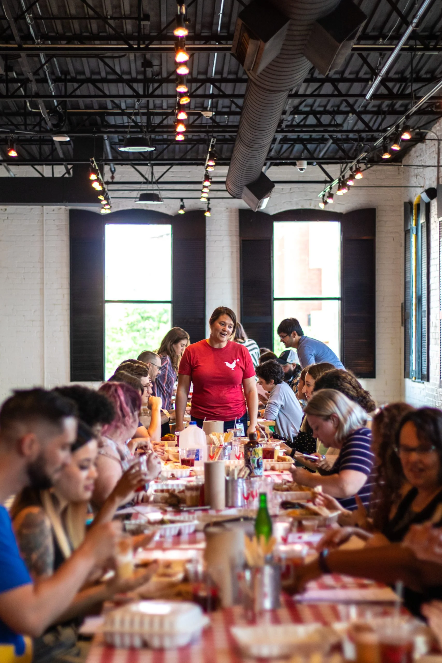 A woman in a red chicken-logo shirt stands at a long table filled with people eating in a restaurant.