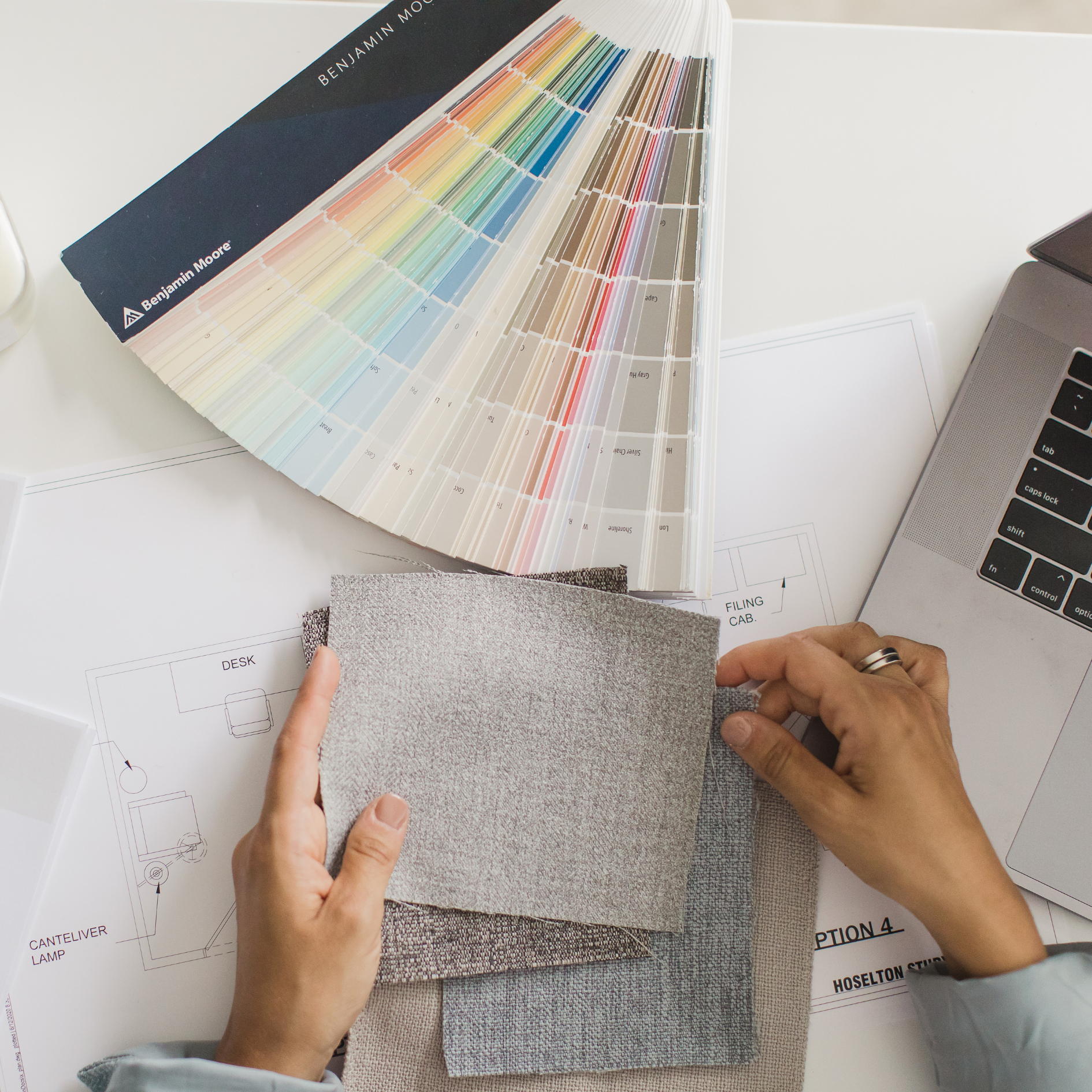 Color swatch sample book open on a table, with pages fanned out displaying various paint colors, alongside architectural and interior design plans, with person holding a fabric sample.