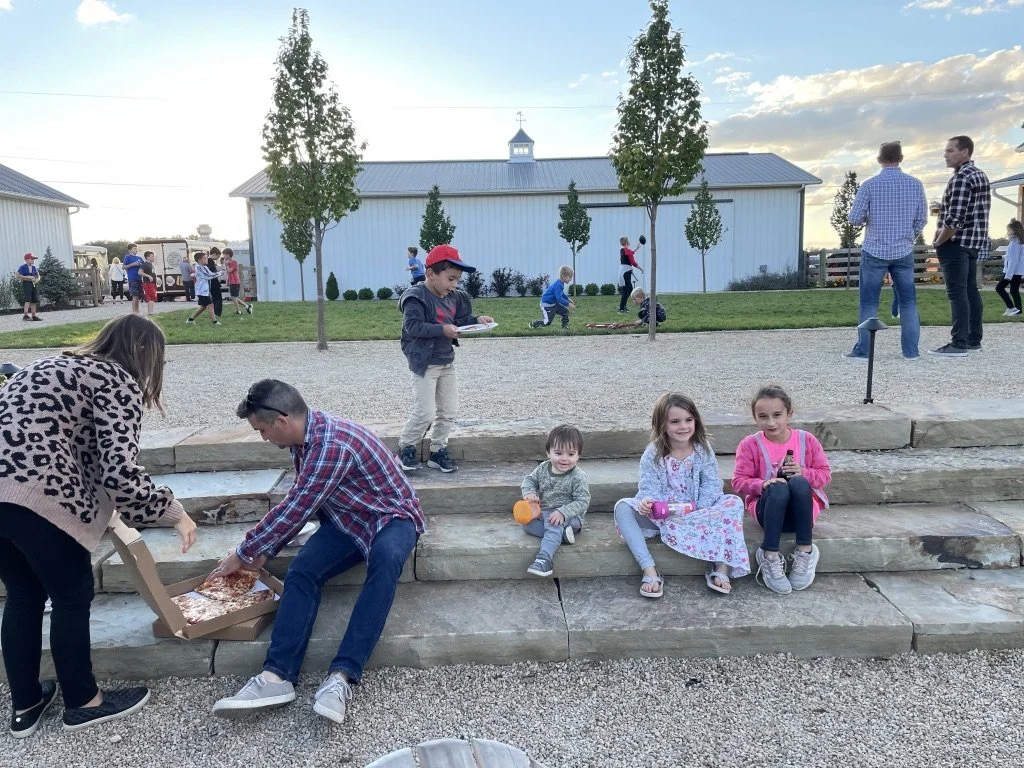 People sitting on stone steps in an outdoor courtyard eating pizza while children play on the grass nearby.