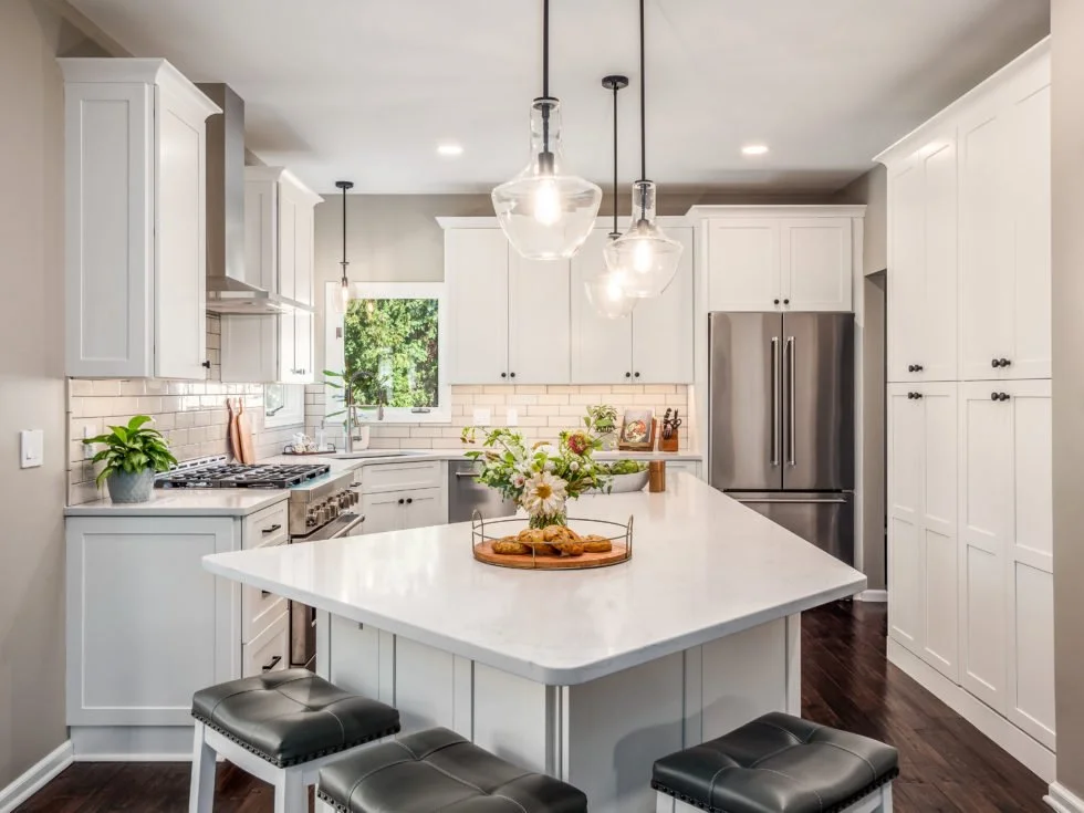 White kitchen with an island, stone countertops, stainless steel appliances, and three glass pendant lights.
