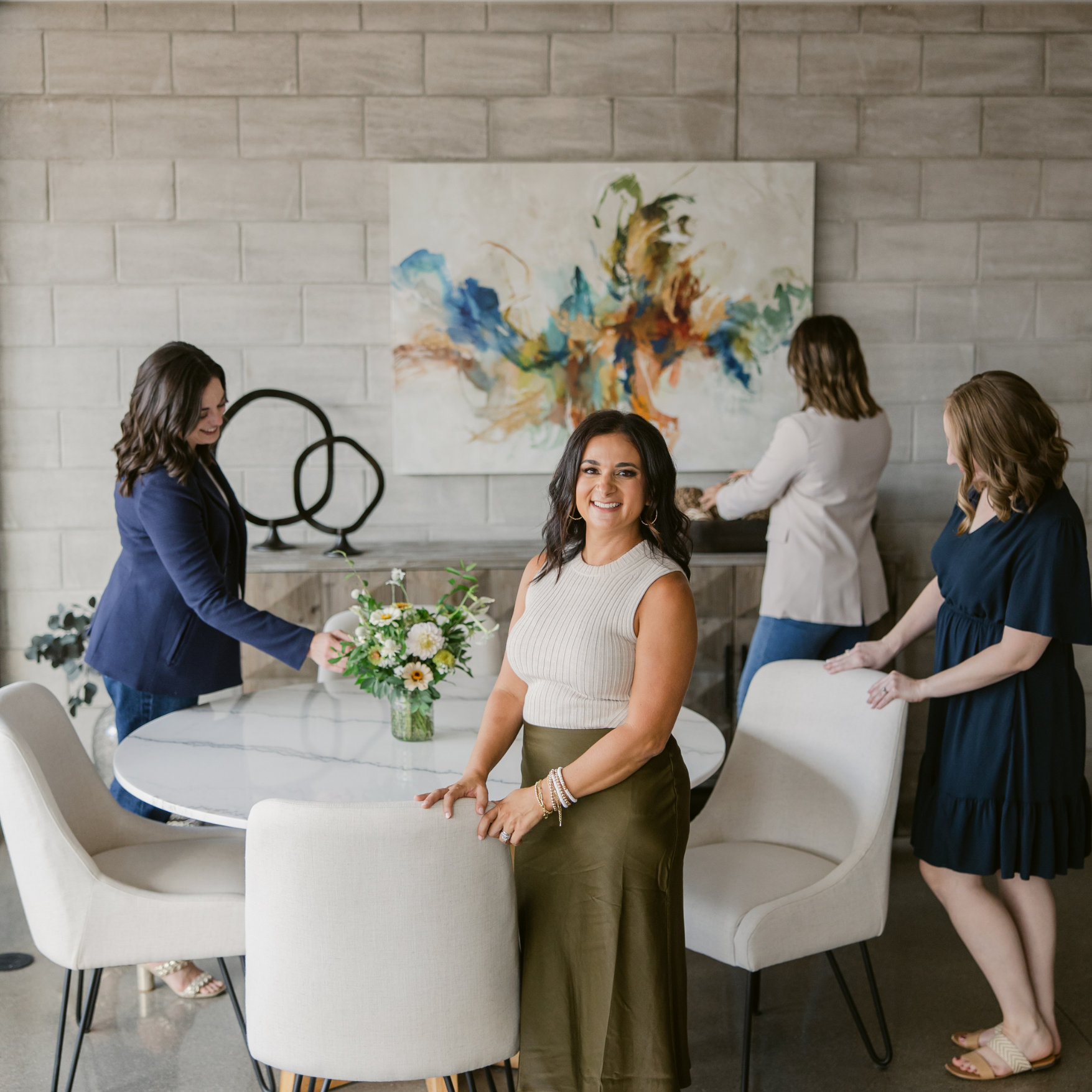 Four women stand in a modern room with a round dining table and an abstract painting.
