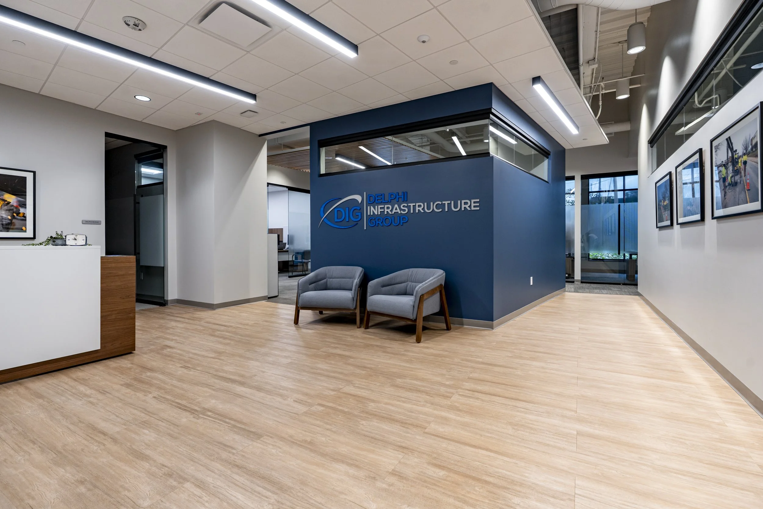 Office lobby area with wooden flooring, a blue wall with the logo 'Delphi Infrastructure Group,' gray chairs, framed photos on the wall, and reception desk visible.