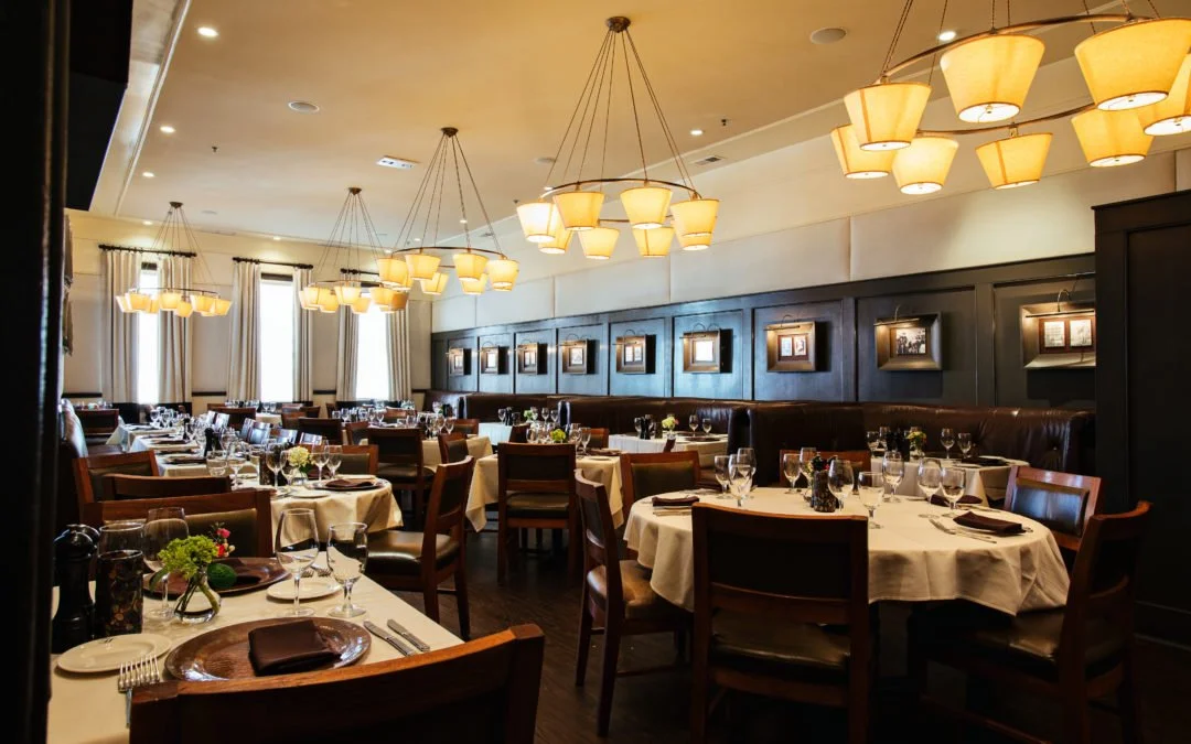 Dining room interior with round tables set for dinner under tiered light fixtures and wood-paneled walls.
