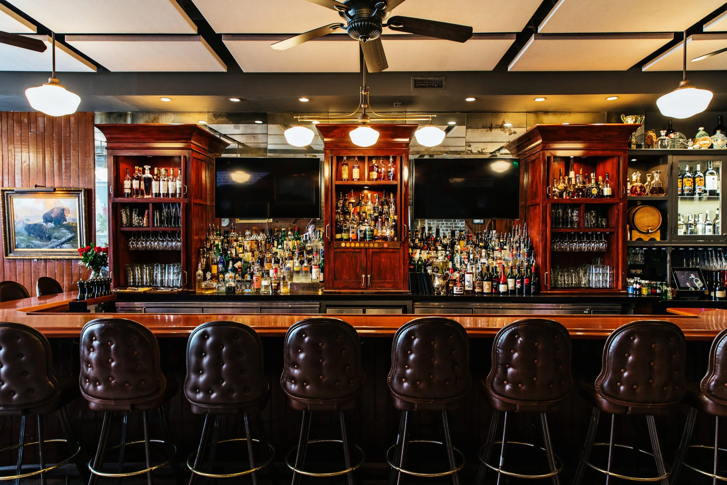 A wooden bar counter with a row of brown leather stools, backed by a large, well-stocked liquor display and two televisions.