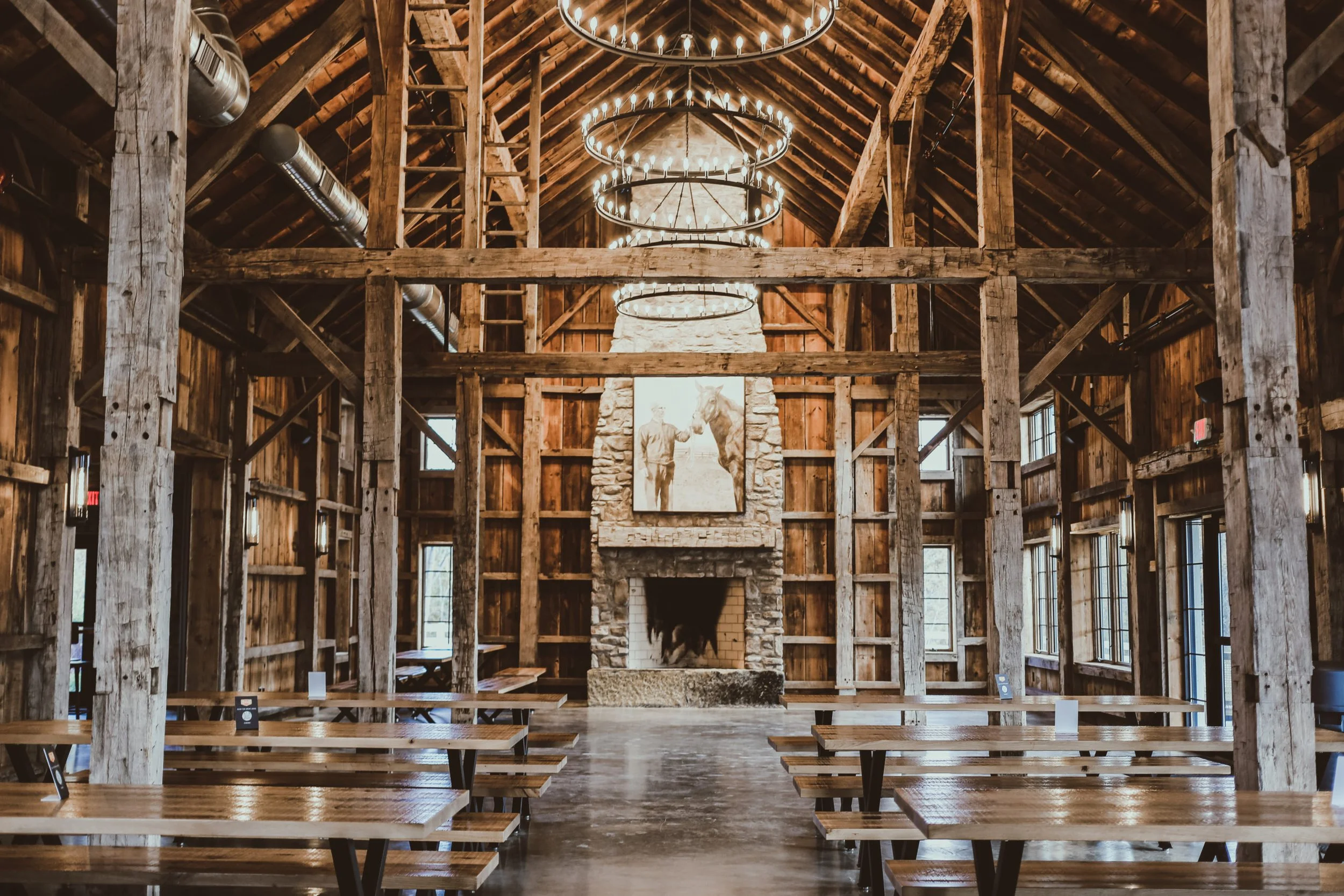 Interior of a rustic wooden barn event space with long tables and two large circular chandeliers hanging above a fireplace.
