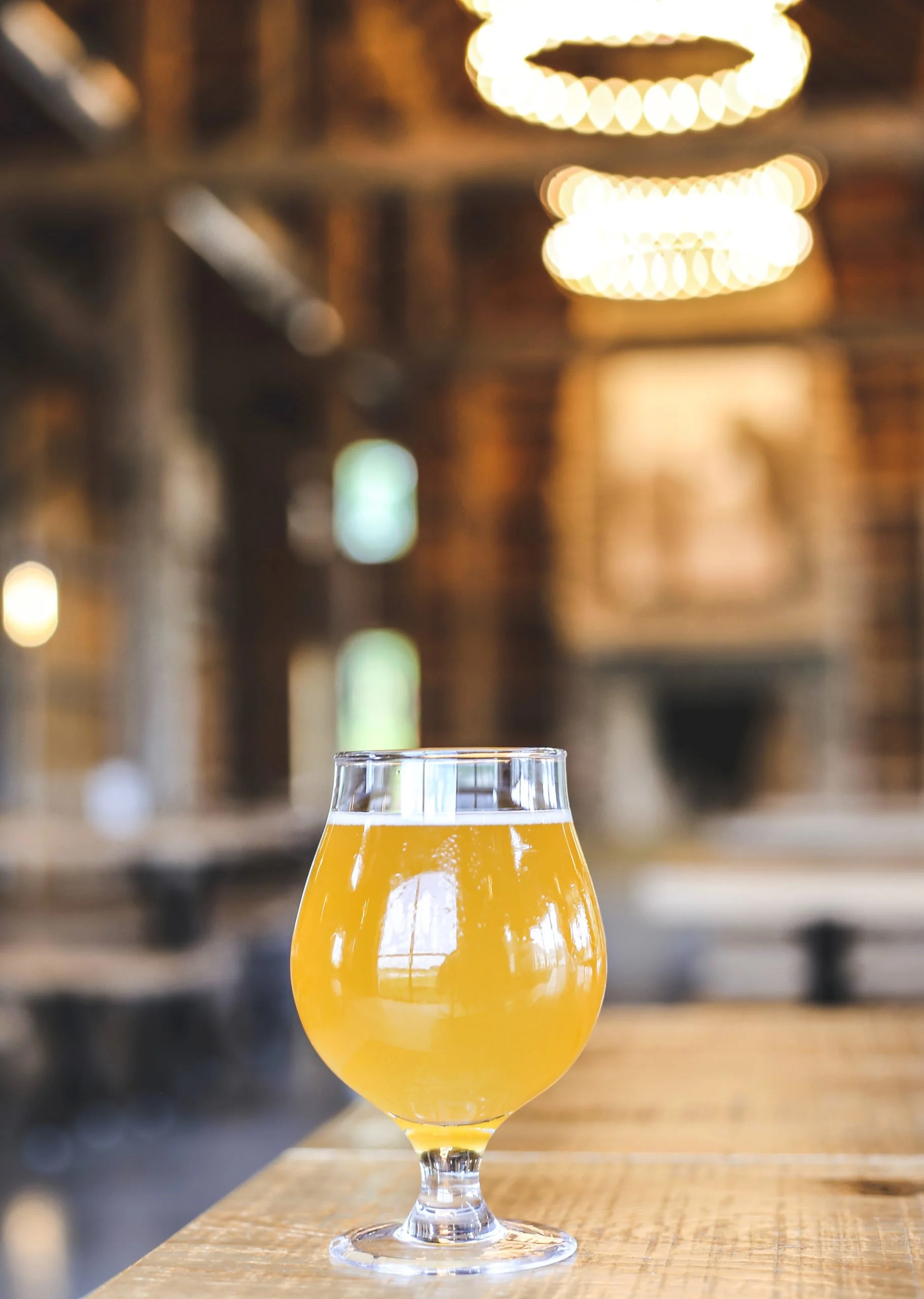 A glass of hazy golden beer on a wooden table inside a rustic building with blurred lights.