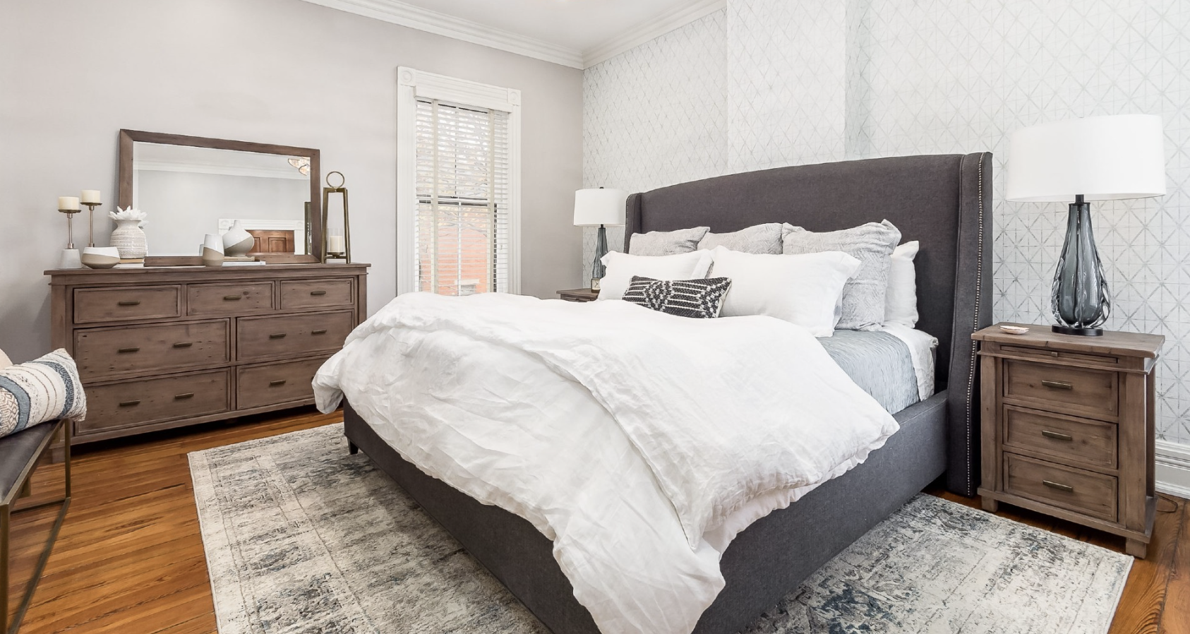 A bedroom featuring a gray upholstered bed, two wooden nightstands with lamps, and a matching dresser with a mirror.