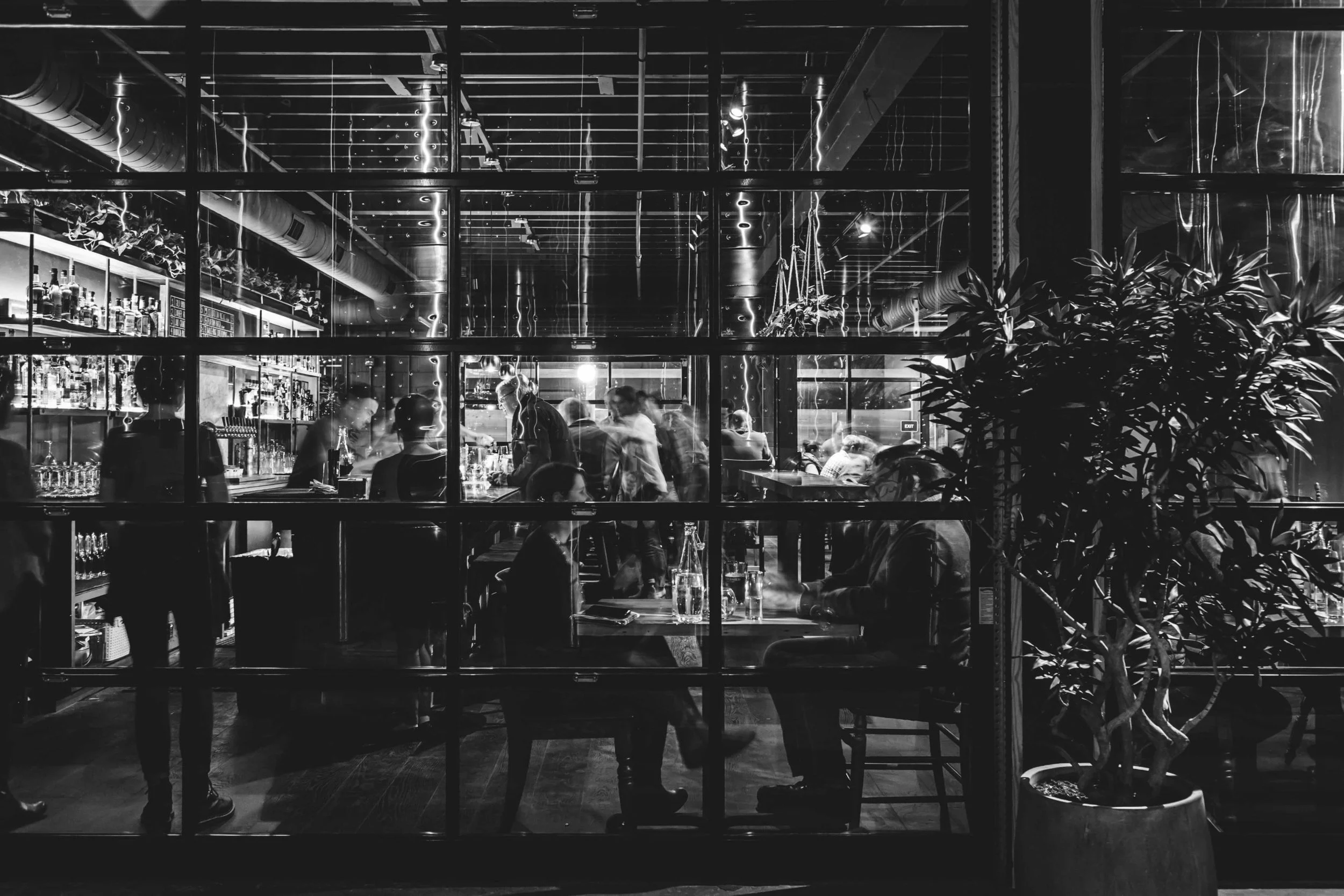 A view through a window into a dimly lit, busy bar with people sitting at tables and the counter.