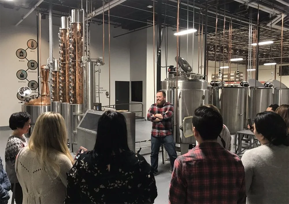 A group of people touring a distillery, listening to a man standing near metal fermentation tanks.
