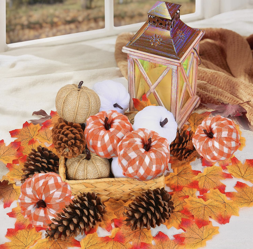 A wicker basket filled with decorative fabric pumpkins, pinecones, and a metal lantern on a bed of fall leaves.