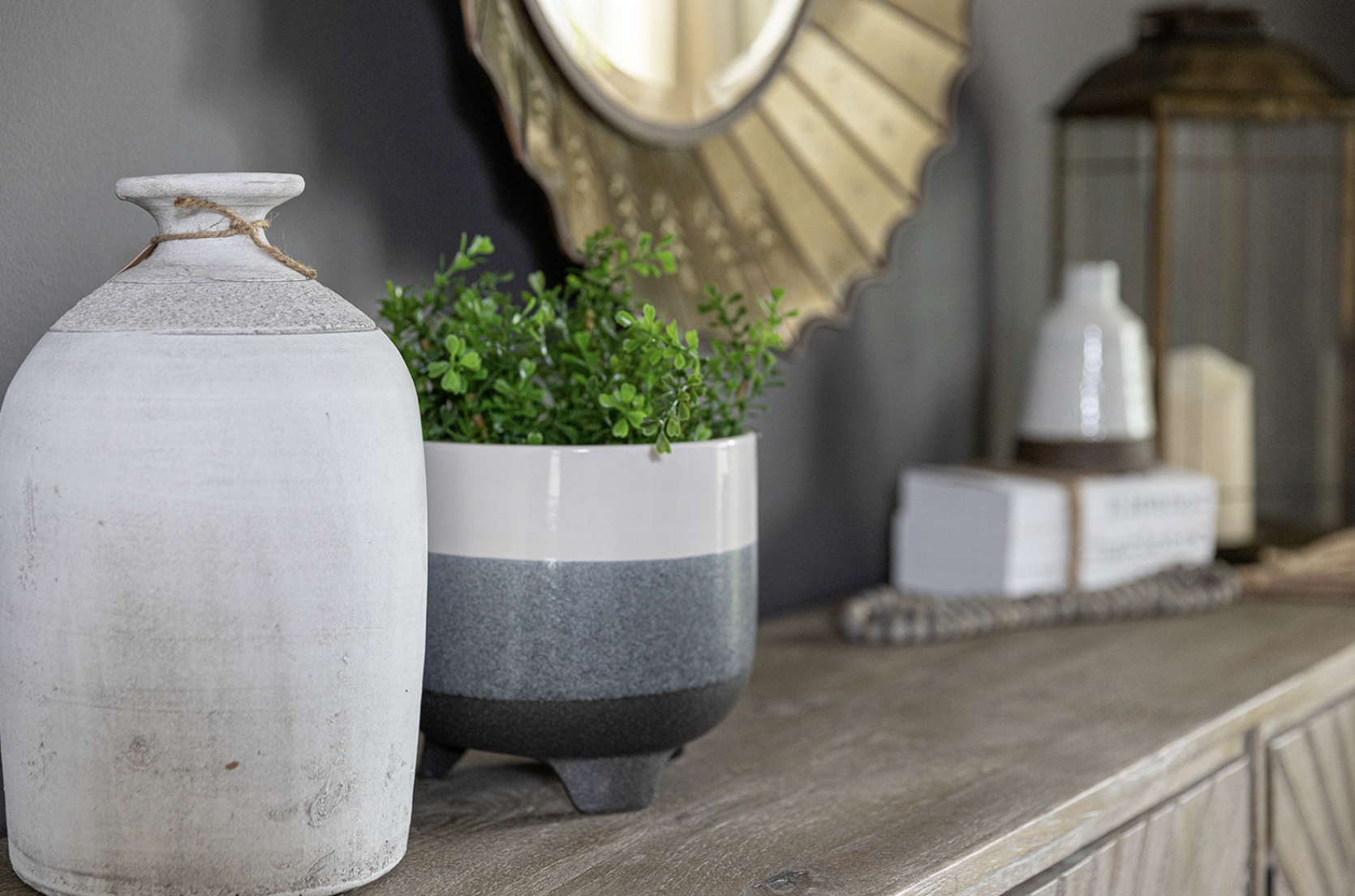 A white textured vase next to a potted green plant in a two-toned gray planter on a wooden surface.