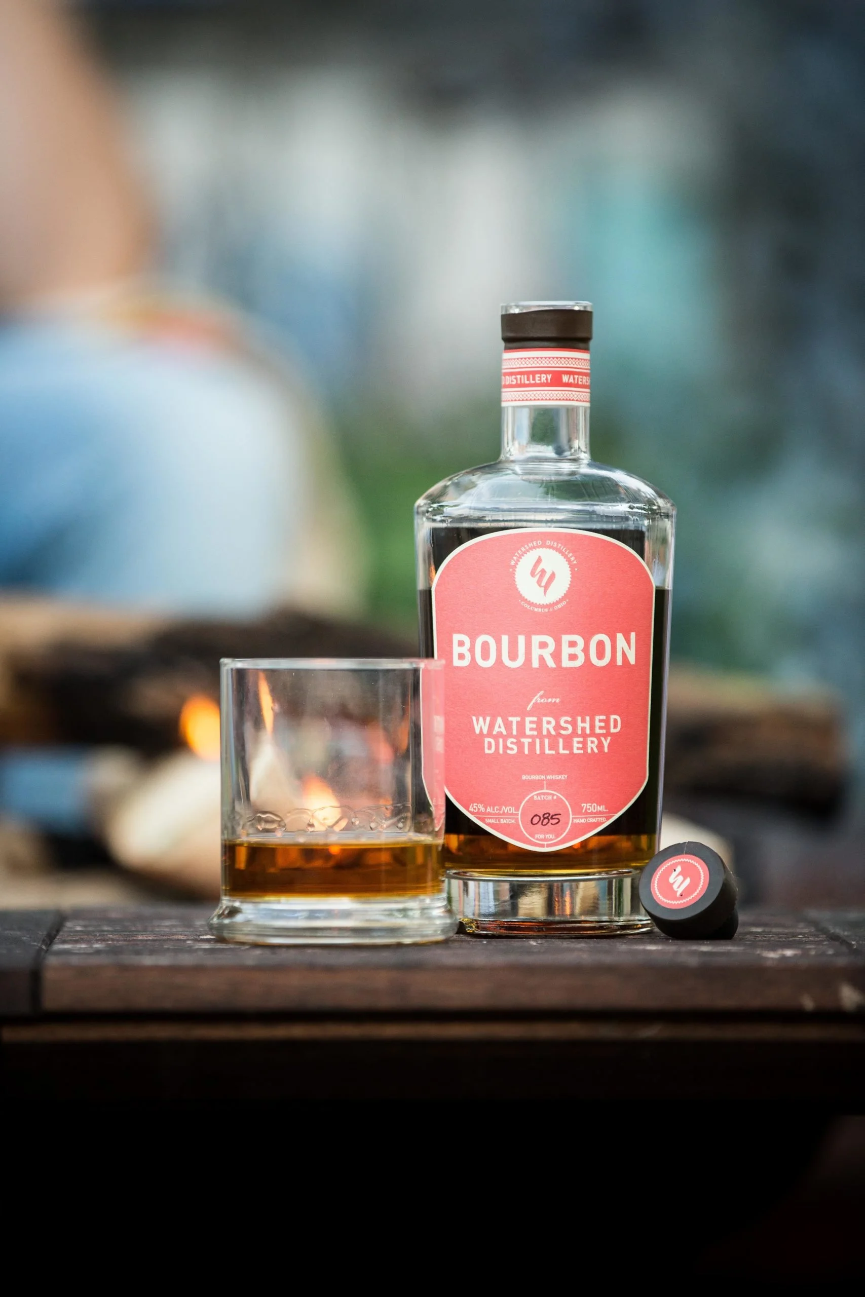 Bottle of Watershed Distillery bourbon and a glass of whiskey on a wooden surface in front of a campfire.