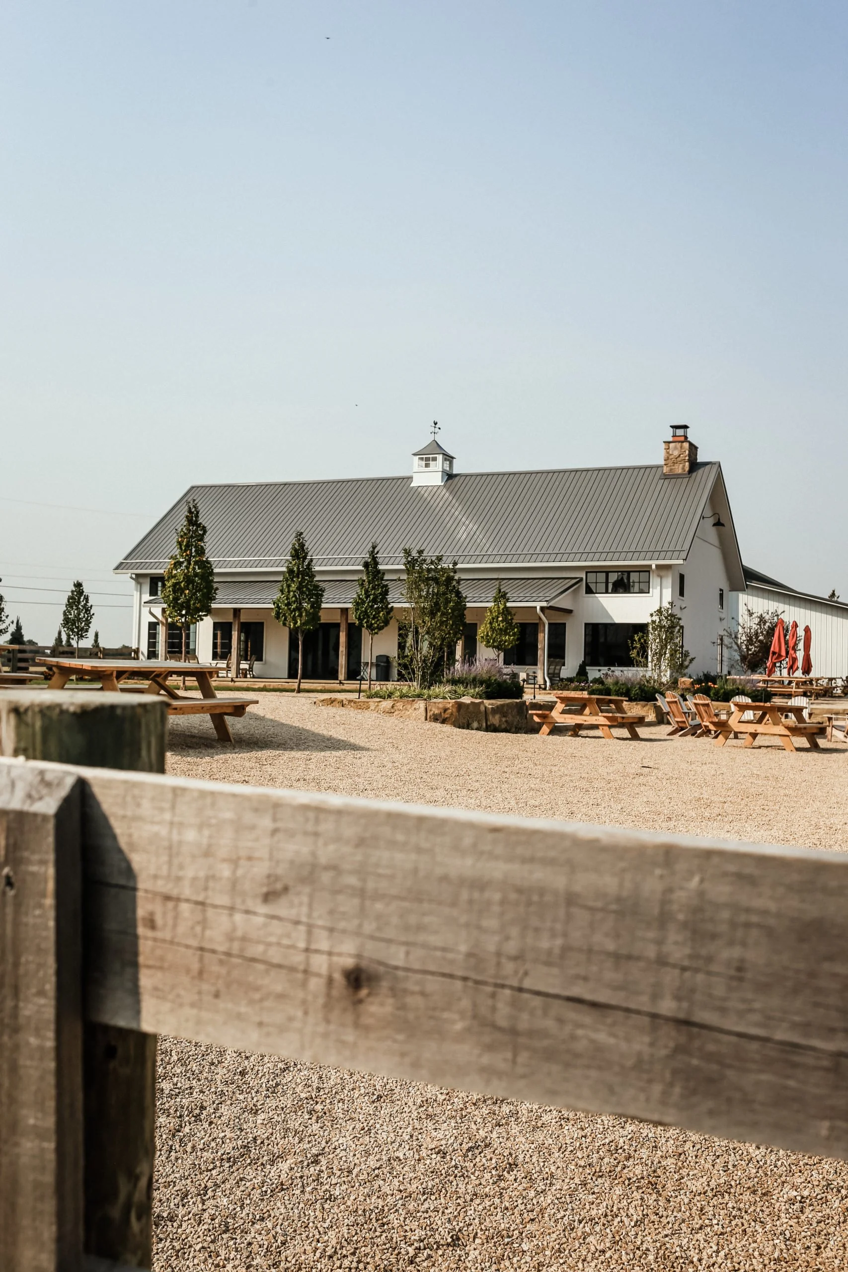 White barn-style building with a metal roof, fronted by a gravel patio with several picnic tables.