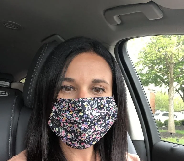 A woman with dark hair sits in a car wearing a floral-patterned face mask.