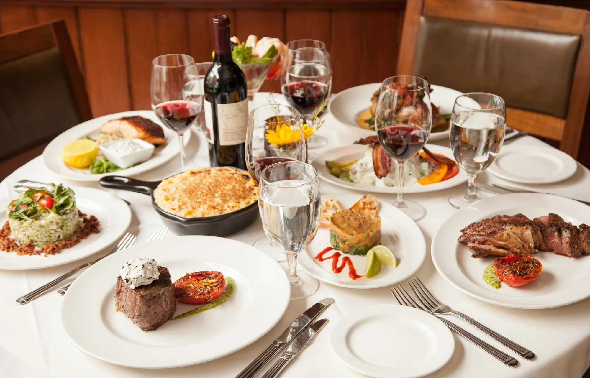 A dinner table set with multiple plates of steak, seafood, sides, and glasses of red wine.