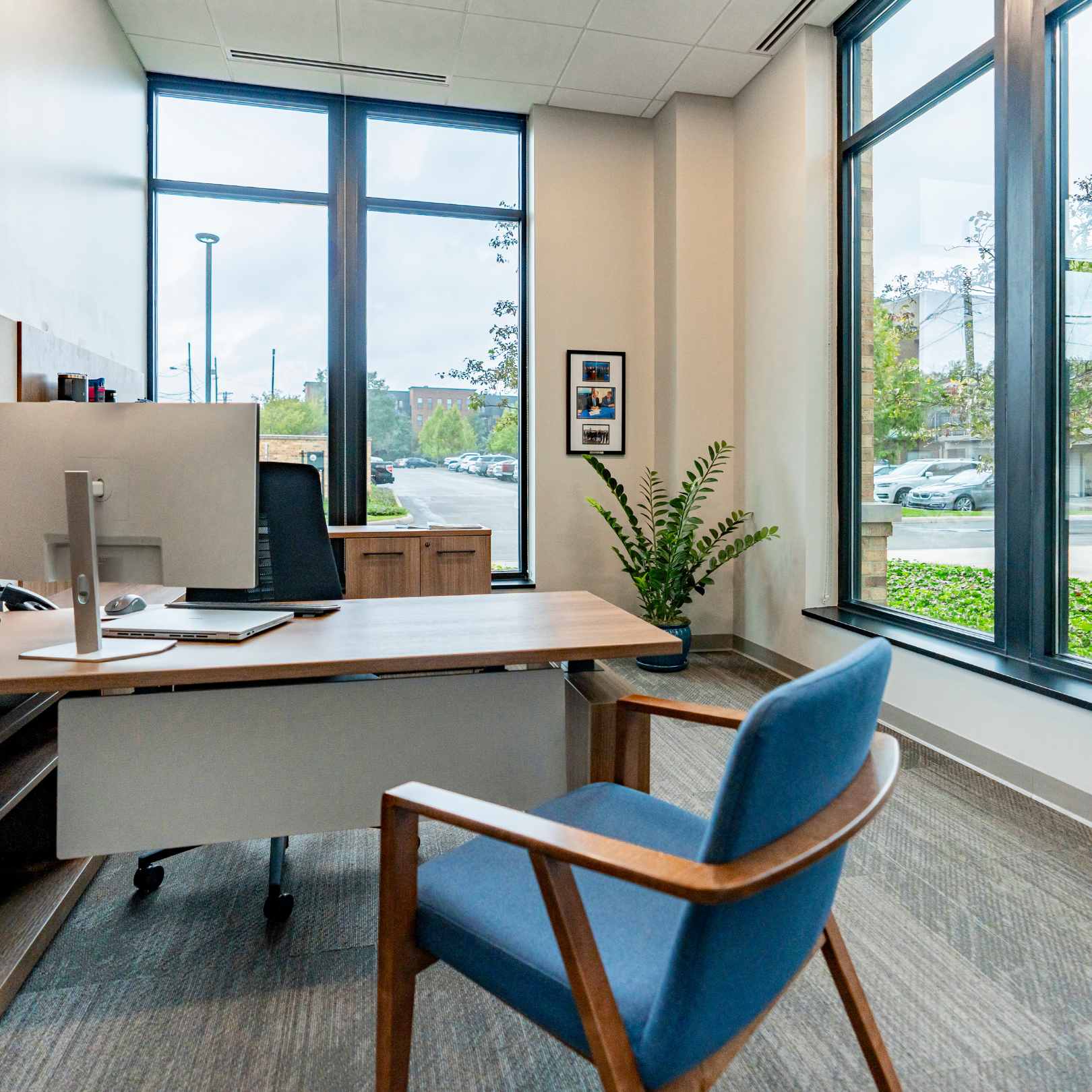An office with a desk, computer, and blue armchair, framed by large windows.