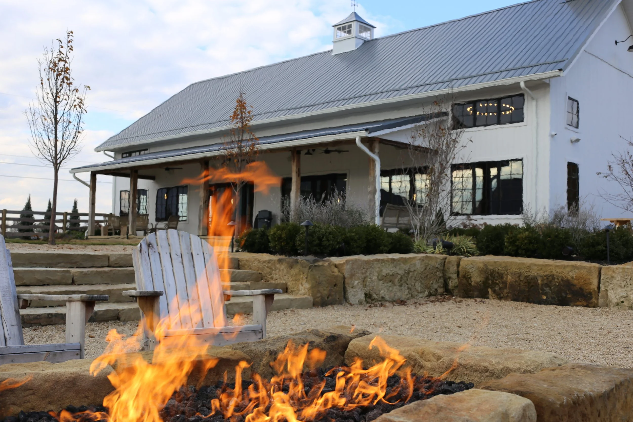Fire pit with flames burning in front of a white barn-style building with a gravel patio and chairs.