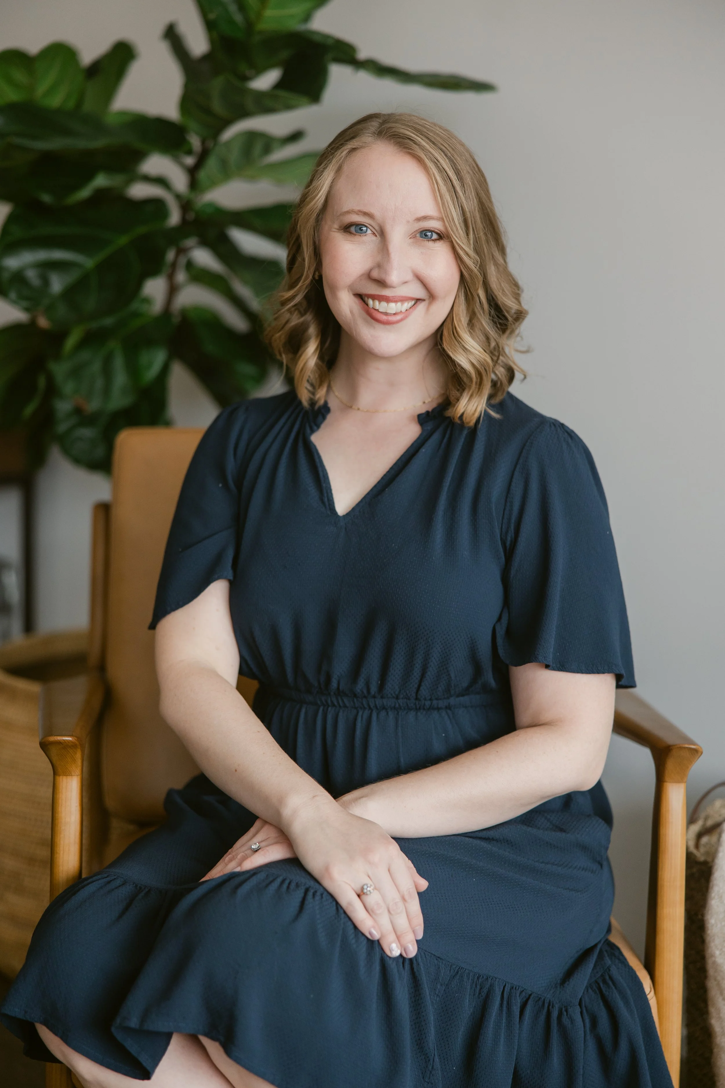 A smiling woman with blonde wavy hair wearing a navy blue dress sitting on a wooden chair. There is a large green leafy plant behind her and a beige wall in the background.