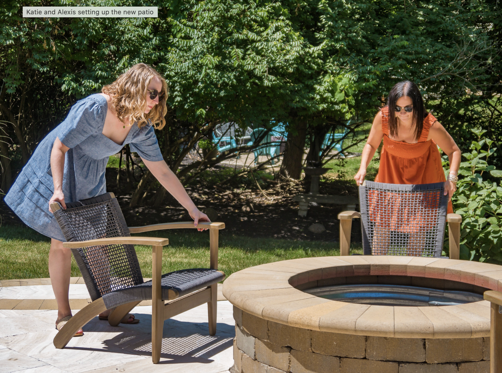 Two women arranging chairs around a circular stone fire pit in a garden.