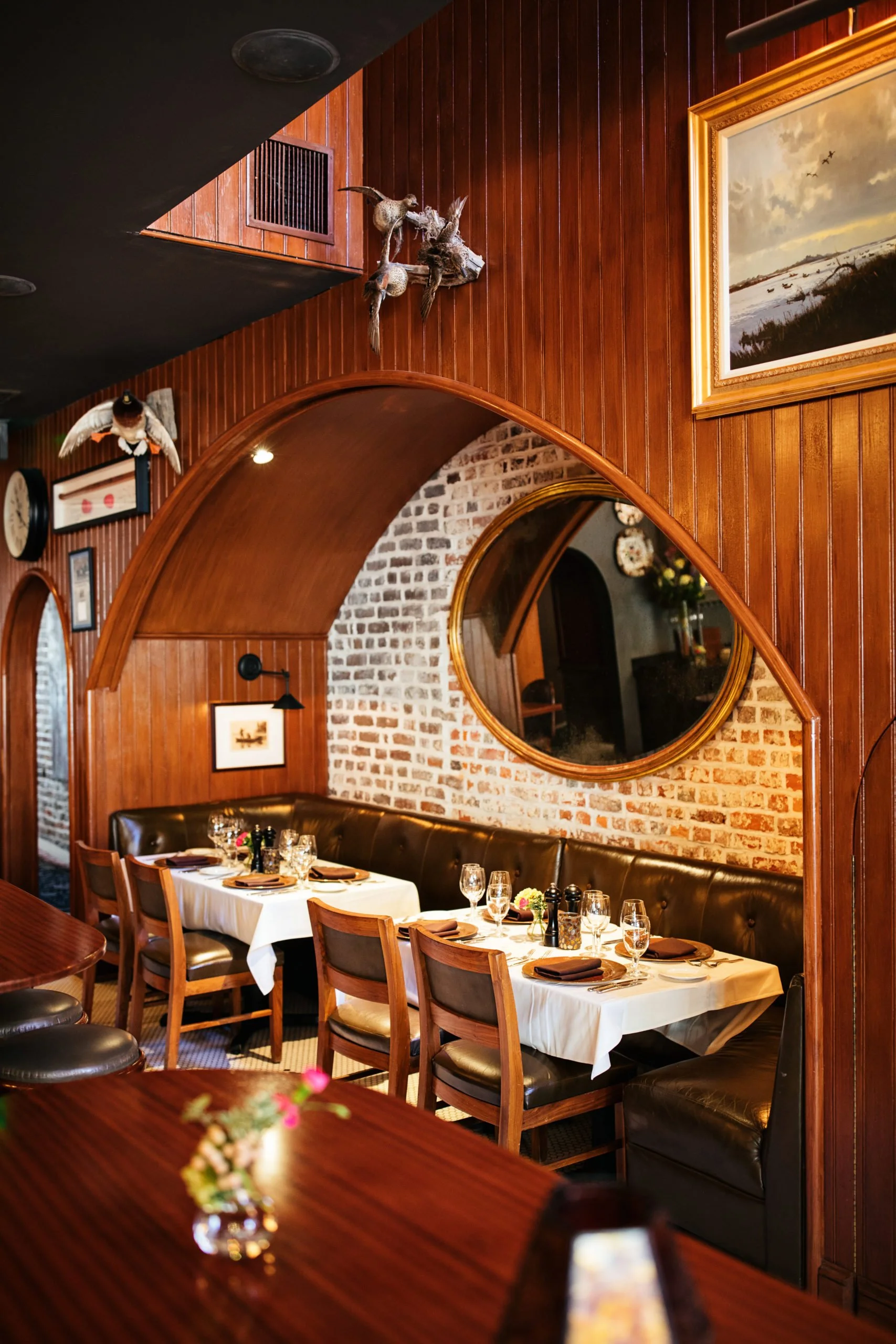 Restaurant interior with leather booths, white-clothed tables, and a large round mirror set into a brick and wood archway.