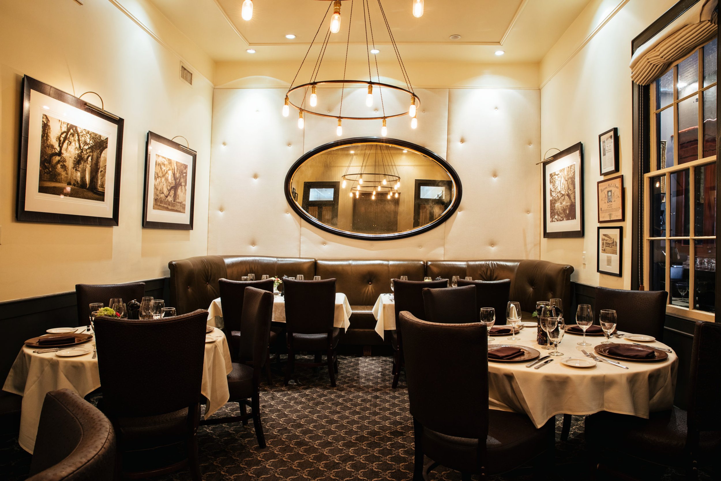 Restaurant interior with round dining tables, leather booth seating, and a large oval mirror on a tufted wall.