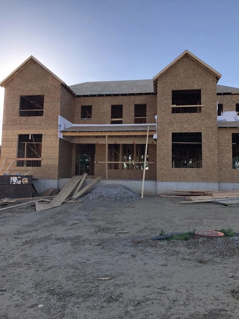 Two-story house under construction with exposed wooden framing and plywood siding.