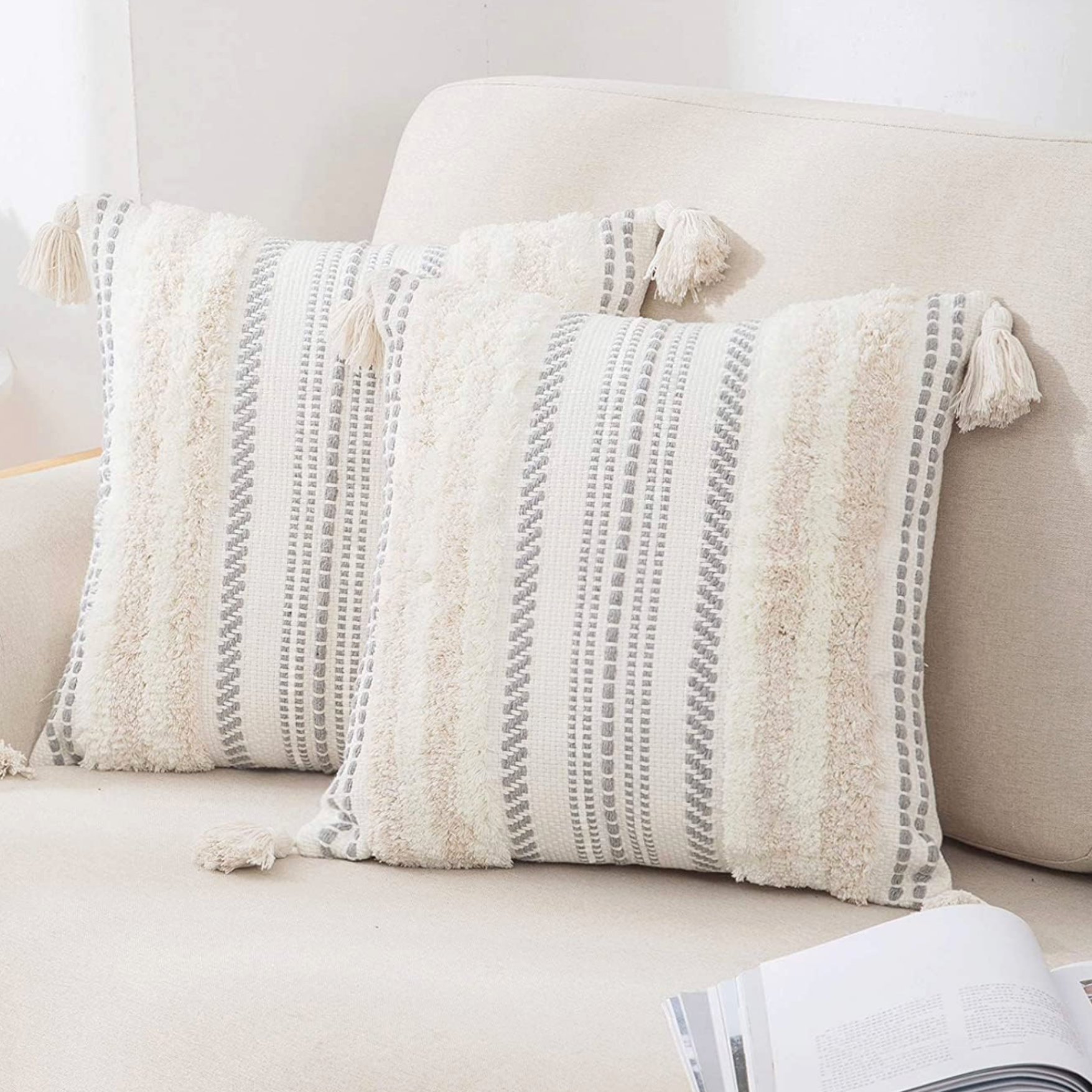 Decorative throw pillows with textured cream and gray striped patterns and tassels on a white sofa, with an open magazine resting on the sofa.