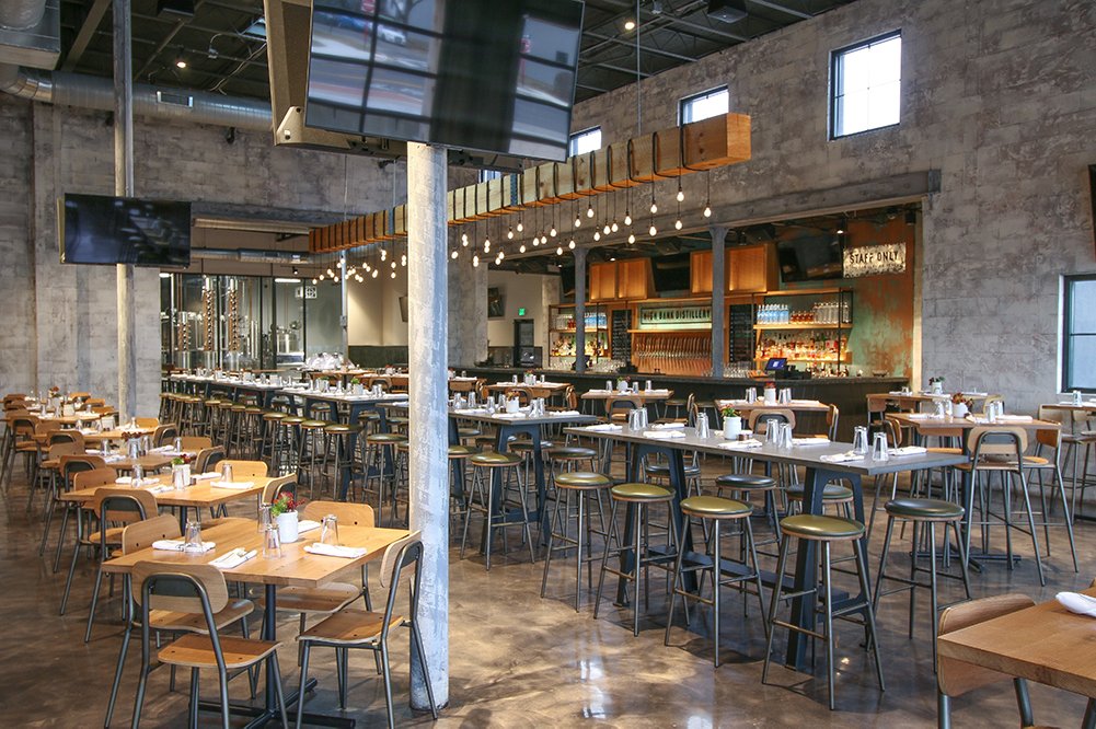 Spacious industrial-style restaurant interior with tables, bar stools, and a long bar in the back.