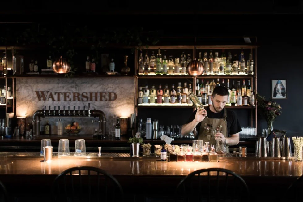 A bartender preparing a drink behind a bar with a “WATERSHED” sign on the back wall.