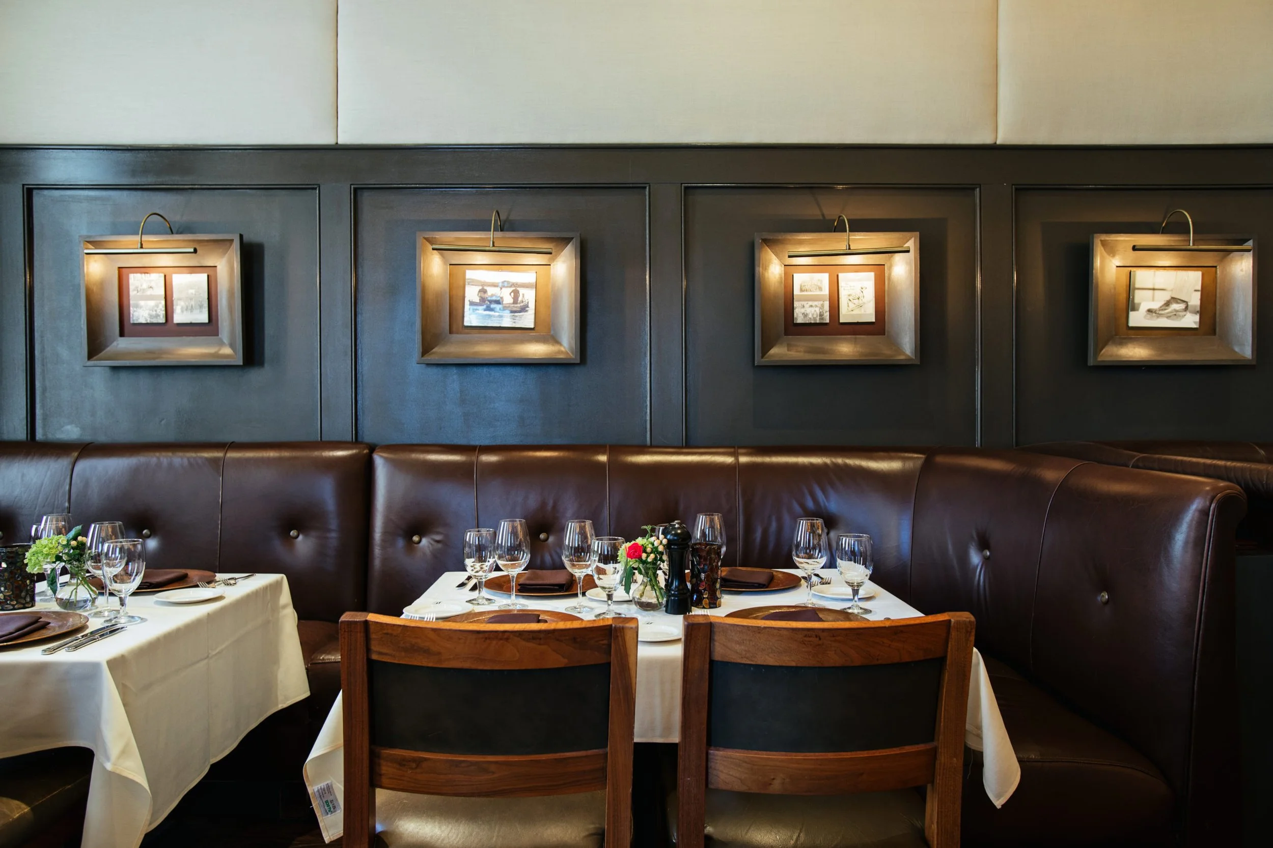 Restaurant booths with brown leather seating, wooden chairs, and tables set with white tablecloths and glassware.
