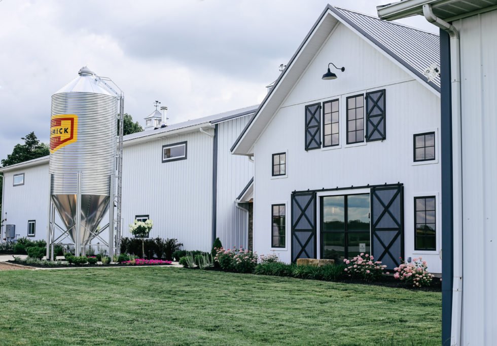 A modern white barn with black shutters and a metal grain silo on a grassy lawn.