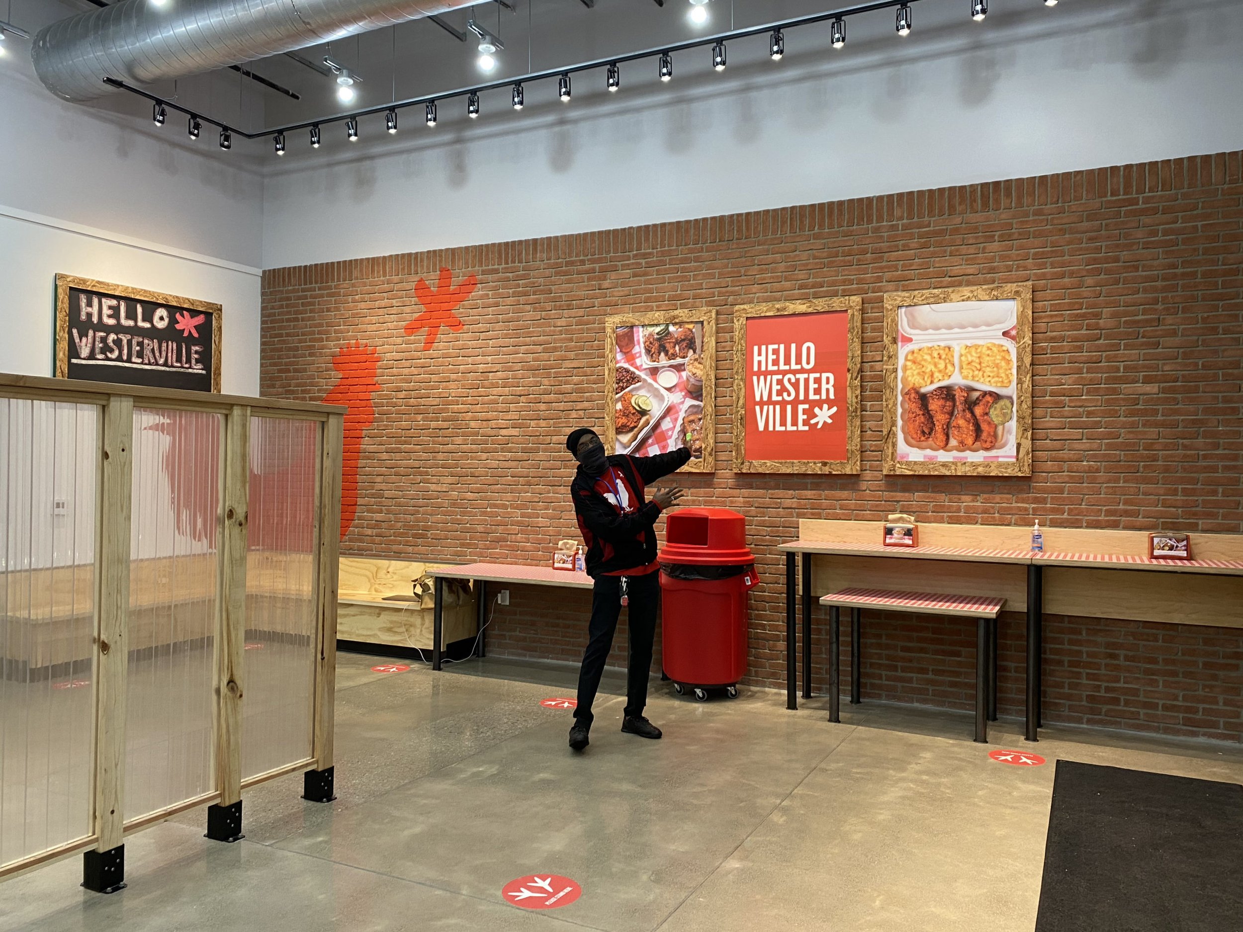 A person in a black jacket and mask stands in a restaurant with brick walls, pointing at framed menus.