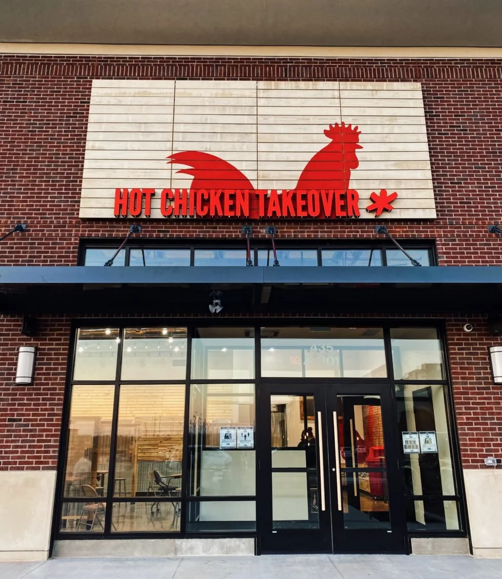 Storefront of Hot Chicken Takeover featuring a large red rooster logo on a wooden panel above glass double doors.