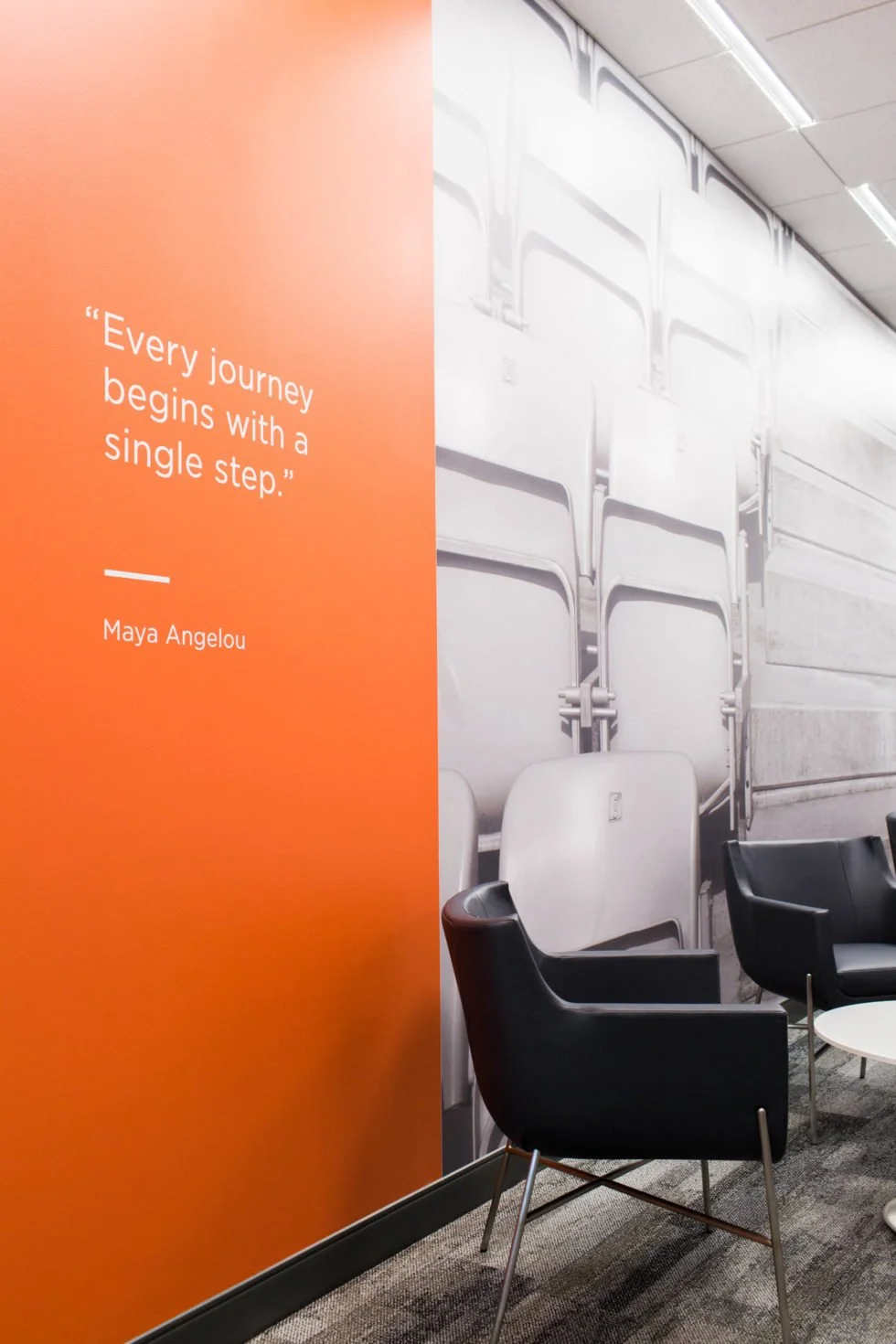 Orange office wall featuring the quote, “Every journey begins with a single step” by Maya Angelou, next to black chairs.