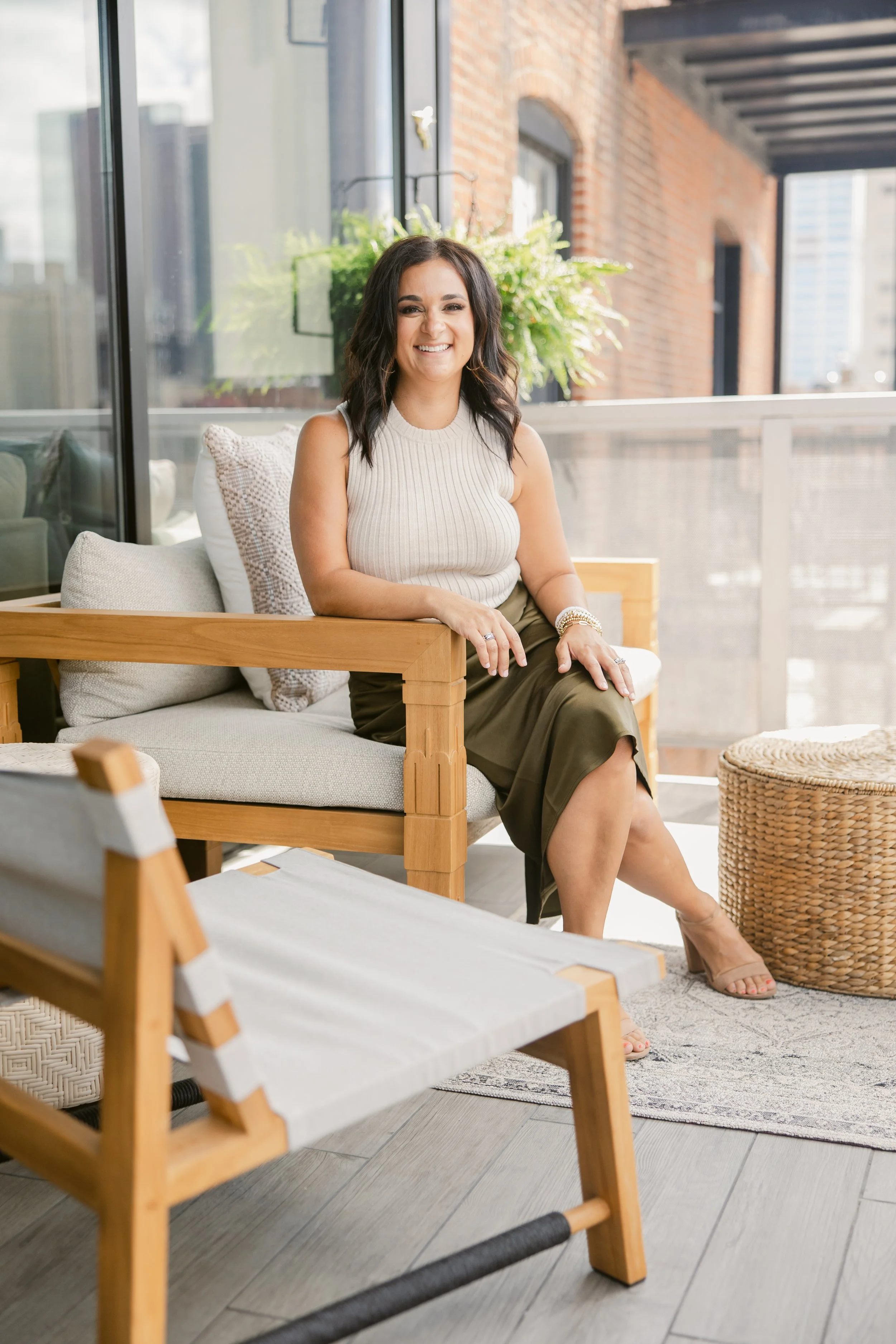 A woman with dark hair, wearing a sleeveless cream top and olive green skirt, sitting on a light-colored couch on a balcony with a glass railing, smiling at the camera. The balcony has plants and brick walls, with a city view in the background.