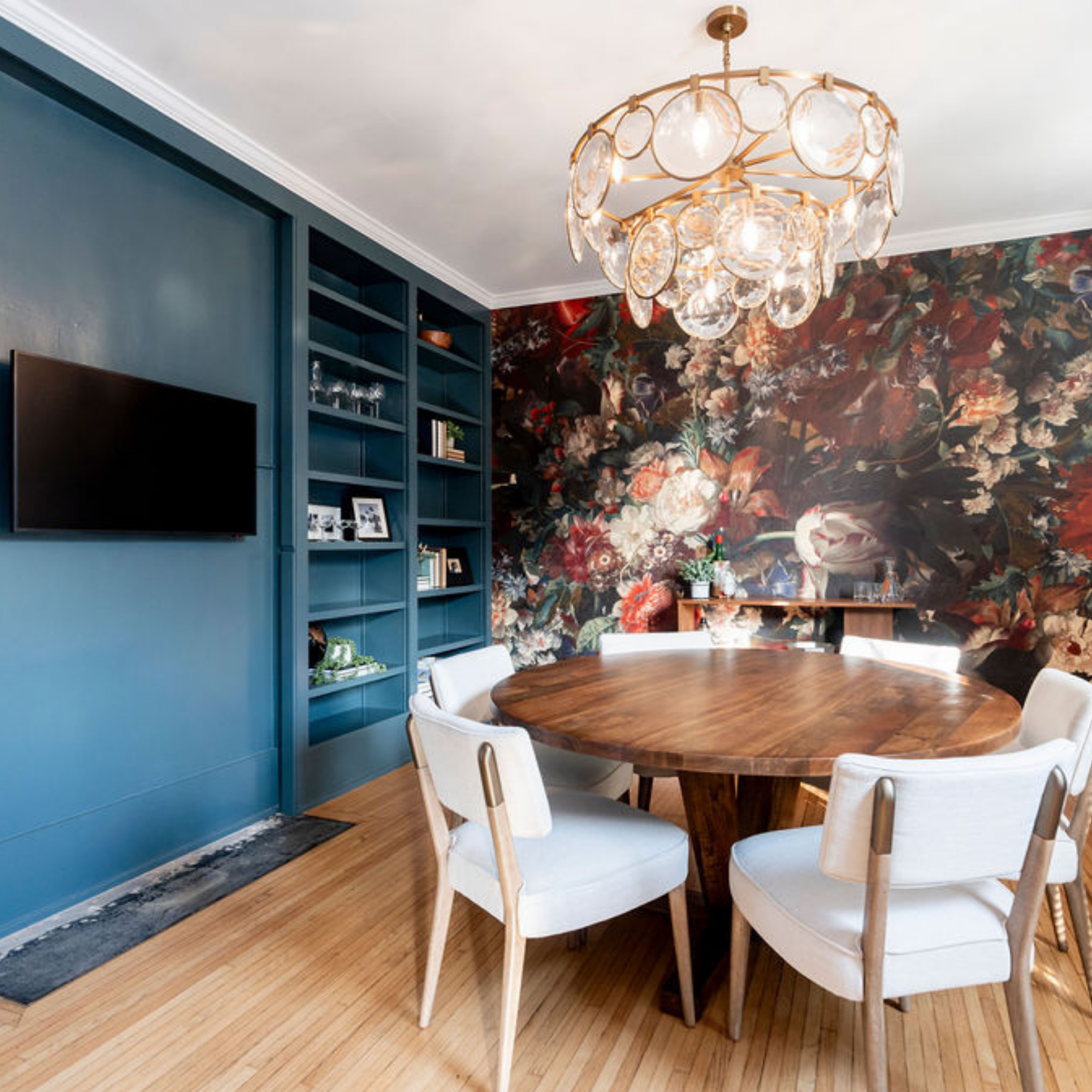 A round wooden dining table with white chairs in front of a dark floral accent wall and blue built-in shelves.