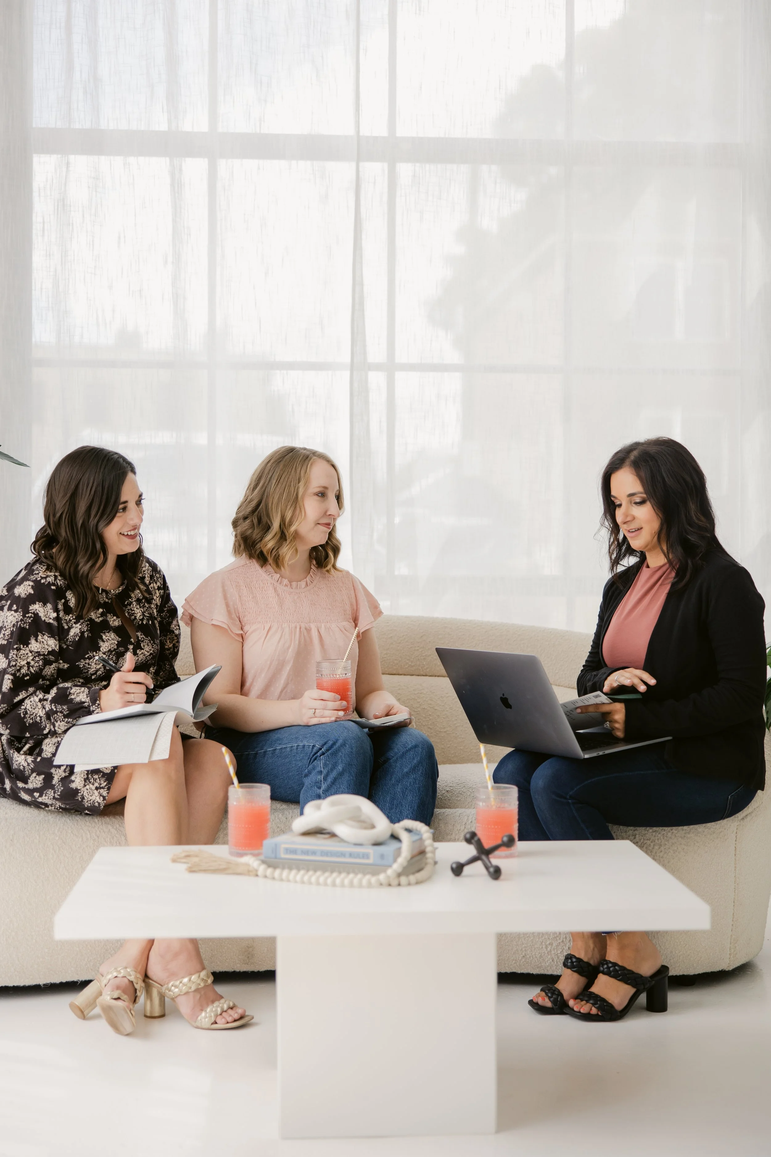 Three women sit on a sofa in a bright room, two reviewing notebooks and one working on a laptop.