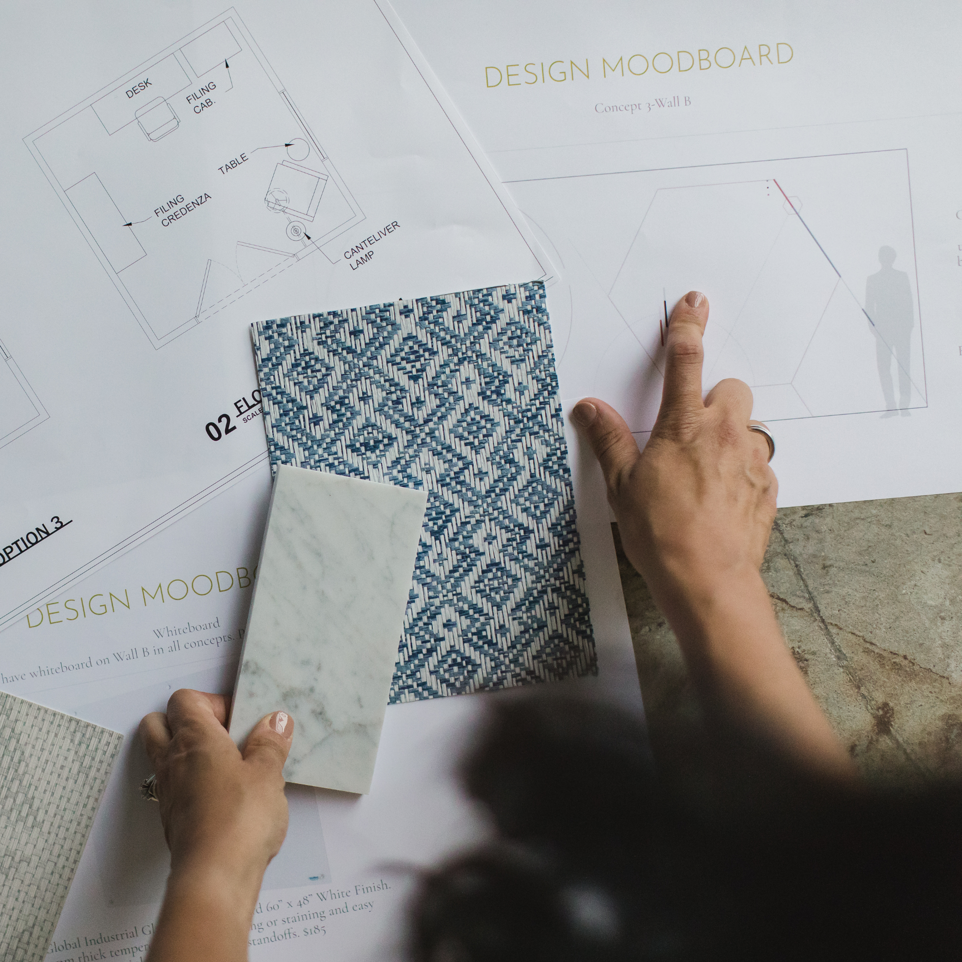 A person is pointing at a design mood board on the table which includes a color and material sample, a detailed architectural floor plan, and printed information about wall design concepts.