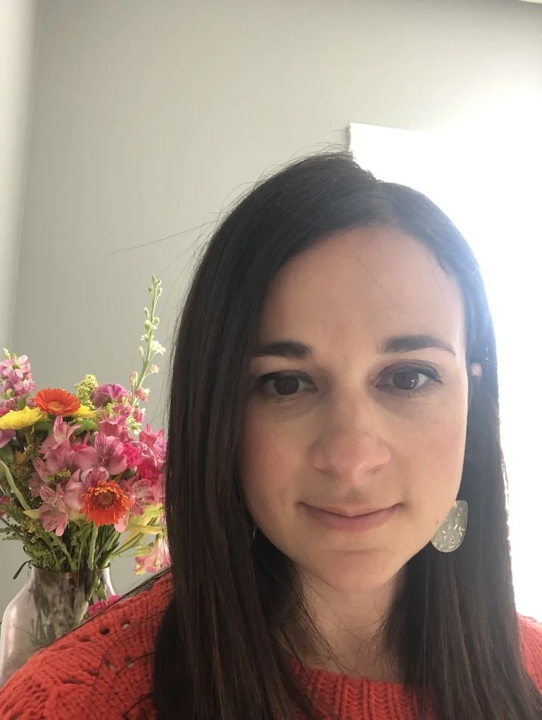 A woman with long dark hair and red sweater stands next to a vase of colorful flowers.